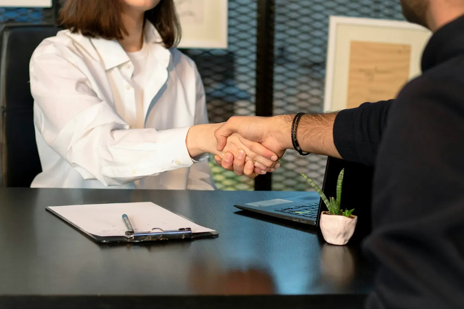 Two people shaking hands across a desk beside a laptop