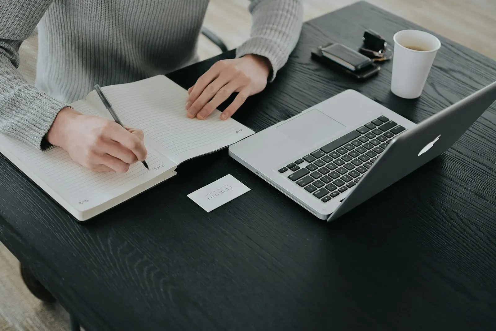 Person writing notes in a notebook beside a laptop and coffee